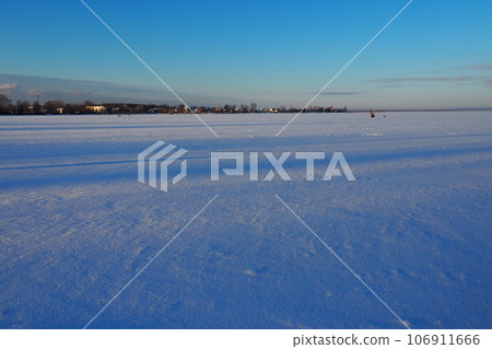 Frozen Lake Logmozero, Karelia, Russia. Winter January in nature. Smooth blue ice surface covered with snow. Evening winter sunset in sunny weather. Horizon with coast. Blue sky Frozen Lake Logmozero, Karelia, Russia. Winter January in nature. Smooth blue ice surface covered with snow. Evening winter sunset in sunny weather. Horizon with coast. Blue sky 106911666
