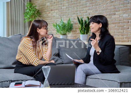 Two business mature women sitting on couch in office, talking, discussing work Two business mature women sitting on couch in office, talking, discussing work 106912421