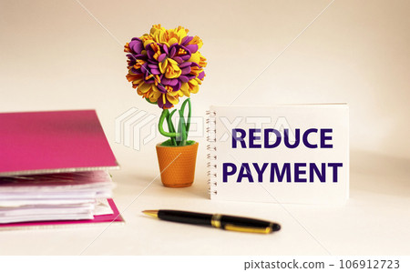 REDUCE PAYMENT text on notepad in front of document folder, pen and flower. 106912723