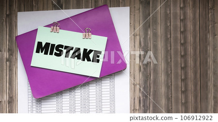 Word MISTAKE, written on a sticker that lies on documents and a wooden table. 106912922