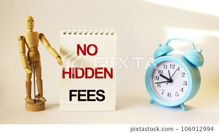 Wooden doll stands near a notepad with the text, No Hidden Fees, near an alarm clock Wooden doll stands near a notepad with the text, No Hidden Fees, near an alarm clock 106912994