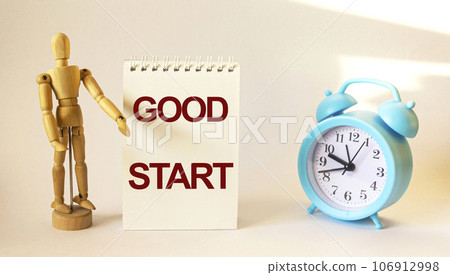 Notepad with the text Good Start on the office table with a wooden doll and an alarm clock. 106912998