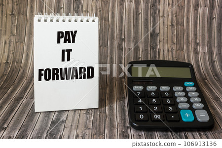 The phrase Pay It Forward with text on a notepad, placed on a wooden board, next to a calculator. The phrase Pay It Forward with text on a notepad, placed on a wooden board, next to a calculator. 106913136