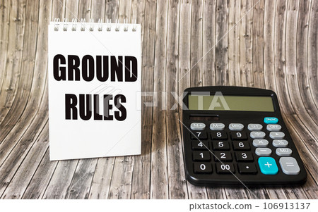 the text GROUND RULES is written on a notebook, next to it lies a caddy calculator on a wooden 106913137