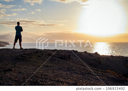 Man looks at the sea and the sunset on the coast of Fuerteventura in the evening light Man looks at the sea and the sunset on the coast of Fuerteventura in the evening light 106915492