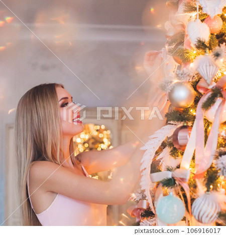 Young cheerful blonde model woman decorating Christmas tree at home. Happy Christmas Holidays Young cheerful blonde model woman decorating Christmas tree at home. Happy Christmas Holidays 106916017