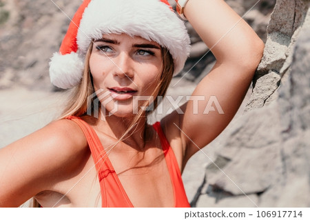 Woman summer travel sea. Happy tourist in red bikini and Santas hat enjoy taking picture outdoors for memories. Woman traveler posing on the beach surrounded by volcanic mountains, sharing travel joy Woman summer travel sea. Happy tourist in red bikini and Santas hat enjoy taking picture outdoors for memories. Woman traveler posing on the beach surrounded by volcanic mountains, sharing travel joy 106917714