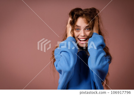 Happy surprised young adult woman wearing winter blue sweater and hold head with her hands looking at camera with joyful smile Happy surprised young adult woman wearing winter blue sweater and hold head with her hands looking at camera with joyful smile 106917796