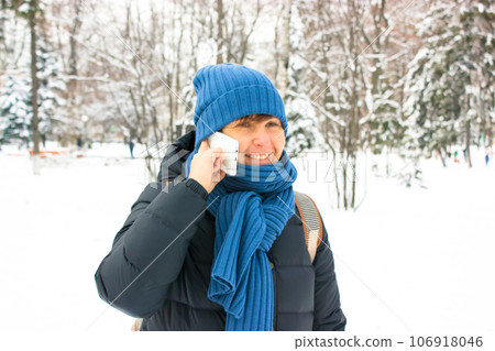 A girl wearing blue scarf hat smiles talking on smartphone in snow-covered park woods in winter. Young European woman of 35-40 years old chatting cheerfully on phone standing in snow-covered landscape 106918046