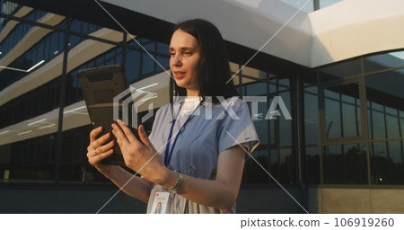Female doctor stands outdoors near hospital, talks to patient using digital tablet 106919260