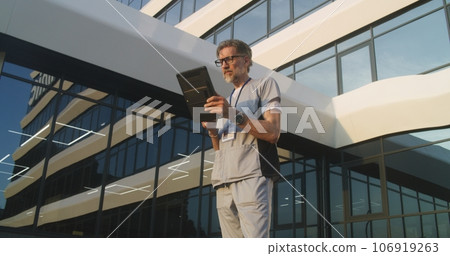 Doctor stands outdoors near clinic, consults patient using digital tablet 106919263