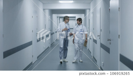 Doctors walk along the clinic corridor, discuss work, use tablet Doctors walk along the clinic corridor, discuss work, use tablet 106919265