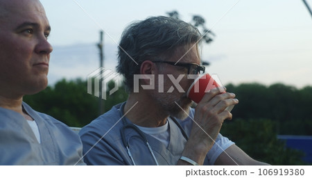 Two doctors sit on bench, drink coffee during break, discuss work 106919380