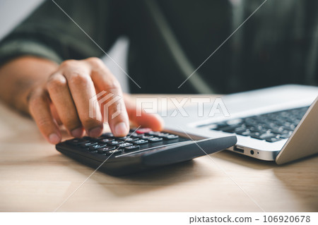 Hand on calculators, aiming for home refinance. Wooden house model, buy or rent note on desk. Concept of saving to buy property, wise mortgage payment. Tax, credit analysis for financial benefit. 106920678