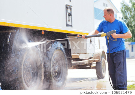Man in overalls washes truck with water gun. Car wash for trucks. Car washer sprays car with Man in overalls washes truck with water gun. Car wash for trucks. Car washer sprays car with 106921479