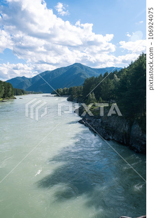 A wide, full-flowing mountain river with a fast current. Large stones stick out of the water. The large turquoise-colored mountain river Katun in the Altai Mountains, Altai Republic. A wide, full-flowing mountain river with a fast current. Large stones stick out of the water. The large turquoise-colored mountain river Katun in the Altai Mountains, Altai Republic. 106924075