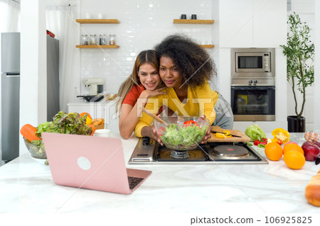 Two women find a moment engrossed in their laptop in the kitchen. A harmonious blend of technology and togetherness, this image depicts the modern age's approach towards collaboration and multitasking Two women find a moment engrossed in their laptop in the kitchen. A harmonious blend of technology and togetherness, this image depicts the modern age's approach towards collaboration and multitasking 106925825