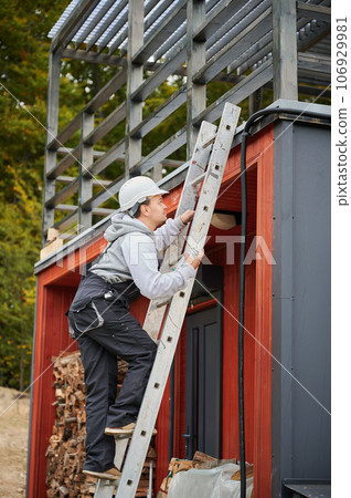 Man worker prepearing for mounting photovoltaic solar modules on roof of house. Electrician in helmet installing solar panel system outdoors. Concept of alternative and renewable energy. 106929981