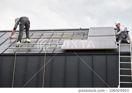 Men measuring photovoltaic solar panels with tape measure. Technicians taking measurements before mounting solar modules on roof of house for generating electricity through photovoltaic effect. 106929983