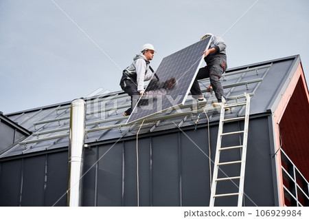 Installers installing solar panel system on roof of house. Men workers in helmets carrying photovoltaic solar module outdoors. Concept of alternative and renewable energy. 106929984