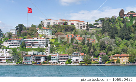 White houses on a green hill by Bosphorus in Istanbul, Turkey, with a large Turkish flag flying overhead White houses on a green hill by Bosphorus in Istanbul, Turkey, with a large Turkish flag flying overhead 106930455