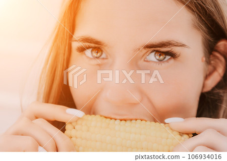 Woman eats corn on beach. Vegetarian hipster woman eat fresh organic grilled corn. Happy lady on sea beach sunset or ocean sunrise. Travel, explore, active yoga and meditation lifestyle concept. 106934016