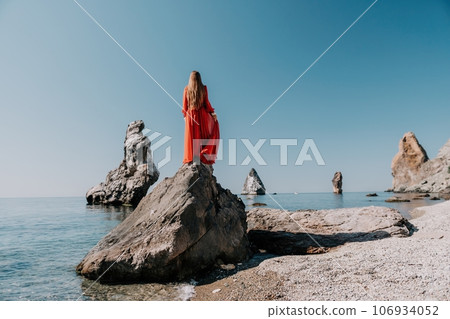 Woman travel sea. Young Happy woman in a long red dress posing on a beach near the sea on background of volcanic rocks, like in Iceland, sharing travel adventure journey Woman travel sea. Young Happy woman in a long red dress posing on a beach near the sea on background of volcanic rocks, like in Iceland, sharing travel adventure journey 106934052