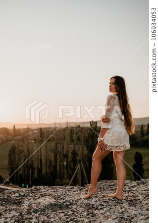 Happy woman in white boho dress on sunset in mountains. Romantic woman with long hair standing with her back on the sunset in nature in summer with open hands. Silhouette. Nature. Sunset. Happy woman in white boho dress on sunset in mountains. Romantic woman with long hair standing with her back on the sunset in nature in summer with open hands. Silhouette. Nature. Sunset. 106934053
