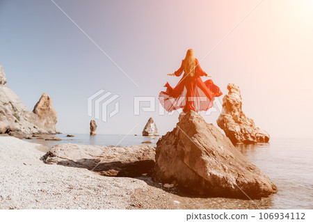 Woman travel sea. Young Happy woman in a long red dress posing on a beach near the sea on background of volcanic rocks, like in Iceland, sharing travel adventure journey Woman travel sea. Young Happy woman in a long red dress posing on a beach near the sea on background of volcanic rocks, like in Iceland, sharing travel adventure journey 106934112