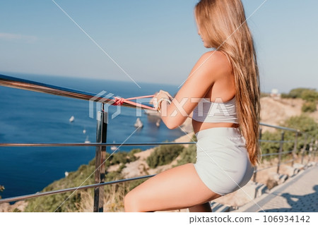 Fitness woman sea. Outdoor workout with fitness rubber bands in park over beach. Female fitness yoga routine concept. Healthy lifestyle. Happy fit woman exercising with rubber band in park. 106934142