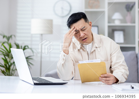 Upset and disappointed man in home office received message mail envelope with bad news, asian man working in home office at desk with laptop. 106934395