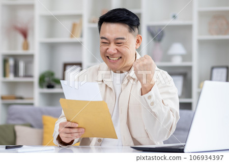 Happy and successful asian received mail notification envelope with good news, man sitting at table at home, working in home office in living room, joyfully celebrating winning news. Happy and successful asian received mail notification envelope with good news, man sitting at table at home, working in home office in living room, joyfully celebrating winning news. 106934397