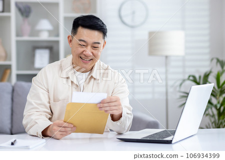 Happy and successful asian received mail notification envelope with good news, man sitting at table at home, working in home office in living room, joyfully celebrating winning news. 106934399
