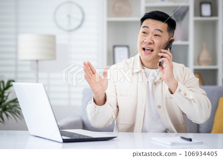 Mature asian man talking on the phone at home, man working in home office sitting at desk in home office, happy and smiling, consulting clients remotely. Mature asian man talking on the phone at home, man working in home office sitting at desk in home office, happy and smiling, consulting clients remotely. 106934405