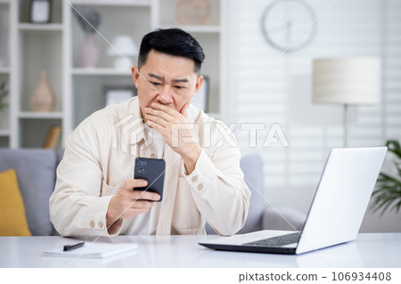 Asian mature man sitting alone at home on sofa in living room cinema, man received online notification message with bad news on phone, upset browsing internet pages using app on smartphone. 106934408