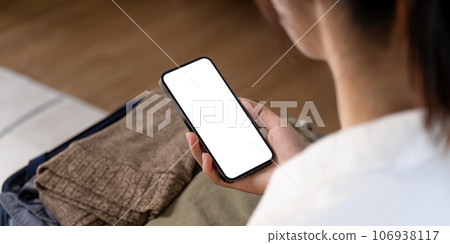 Woman using mobile phone blank white screen display frameless modern design while pack clothes a suitcase ready for travel Woman using mobile phone blank white screen display frameless modern design while pack clothes a suitcase ready for travel 106938117