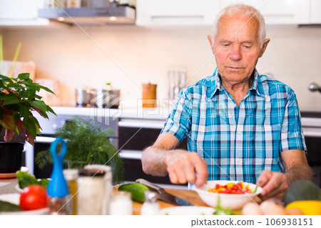 Elderly man cuts vegetables for salad at the table in the kitchen Elderly man cuts vegetables for salad at the table in the kitchen 106938151