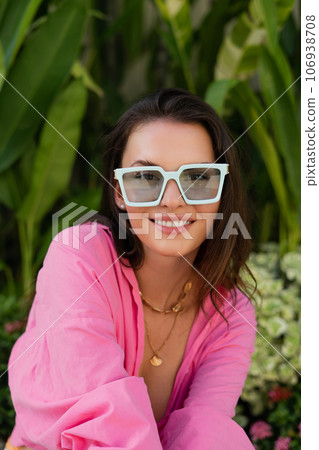 Young beautiful brunette in a pink shirt, neck jewelry, necklace, trendy sunglasses sits on a background of tropical leaves, laughs, smiles Young beautiful brunette in a pink shirt, neck jewelry, necklace, trendy sunglasses sits on a background of tropical leaves, laughs, smiles 106938708