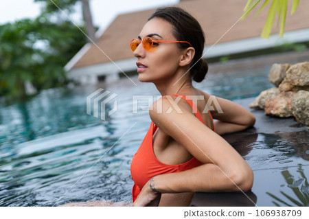 Beautiful european women in two pieces orange bikini swimsuit, tanned, with trendy sunglasses outside at tropical swimming pool Beautiful european women in two pieces orange bikini swimsuit, tanned, with trendy sunglasses outside at tropical swimming pool 106938709