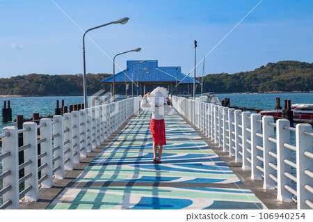 A young slim woman in a red skirt and a white hat stands with her back on a beautiful pier in the andaman sea at rawai beach, phuket, thailand A young slim woman in a red skirt and a white hat stands with her back on a beautiful pier in the andaman sea at rawai beach, phuket, thailand 106940254