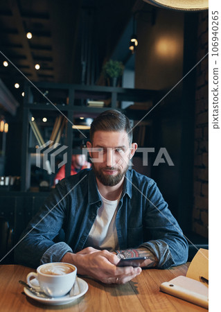 Young bearded businessman,dressed in a denim shirt, sitting at table in cafe and use smartphone. Man using gadget. Young bearded businessman,dressed in a denim shirt, sitting at table in cafe and use smartphone. Man using gadget. 106940265