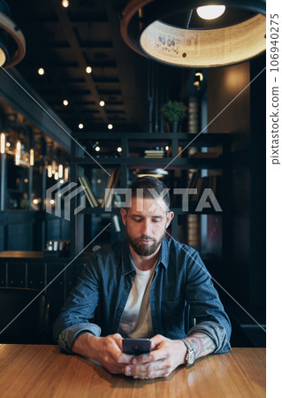 Young bearded businessman,dressed in a denim shirt, sitting at table in cafe and use smartphone. Man using gadget. 106940275