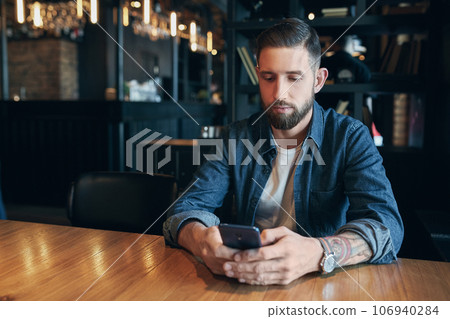 Young bearded businessman,dressed in a denim shirt, sitting at table in cafe and use smartphone. Man using gadget. Young bearded businessman,dressed in a denim shirt, sitting at table in cafe and use smartphone. Man using gadget. 106940284