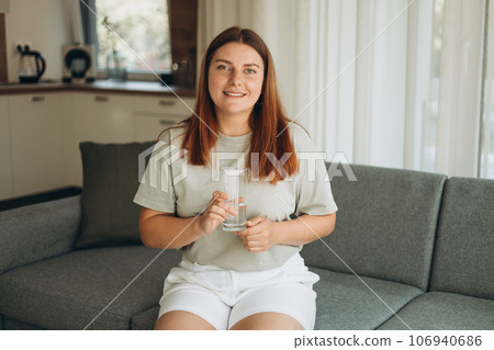 Young woman drinking pure water from glass and sitting on textile couch at home. Health care concept photo, lifestyle, close up 106940686