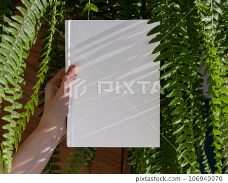 Hands holding book in hardcover mock up among green fern leaf plant Hands holding book in hardcover mock up among green fern leaf plant 106940970
