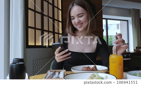 The girl smiles while looking at the phone. A young woman is having dinner in a restaurant, drinking orange juice and eating a steak. Beautiful Woman has lunch in a cafe, lunch in a restaurant. 106941551