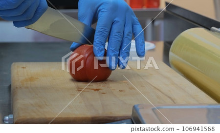 The chef cuts a tomato for a salad, a tomato on a wooden board, a chef in gloves cuts a tomato, a cook in the kitchen prepares ingredients for a salad. 106941568