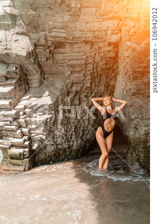 Woman travel sea. Attractive blonde woman in a black swimsuit enjoying the sea air on the seashore around the rocks. Travel and vacation concept. 106942012
