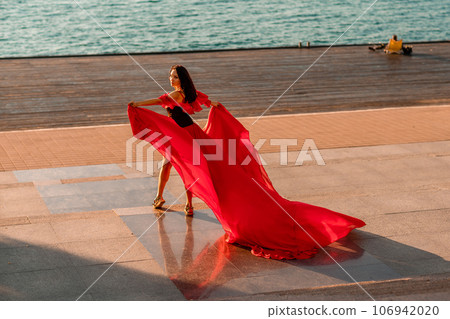 Sunrise red dress. A woman in a long red dress against the backdrop of sunrise, bright golden light of the sun's rays. The concept of femininity, harmony. 106942020