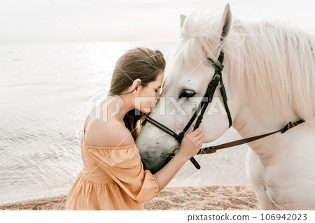 A woman in a dress stands next to a white horse on a beach, with the blue sky and sea in the background. A woman in a dress stands next to a white horse on a beach, with the blue sky and sea in the background. 106942023